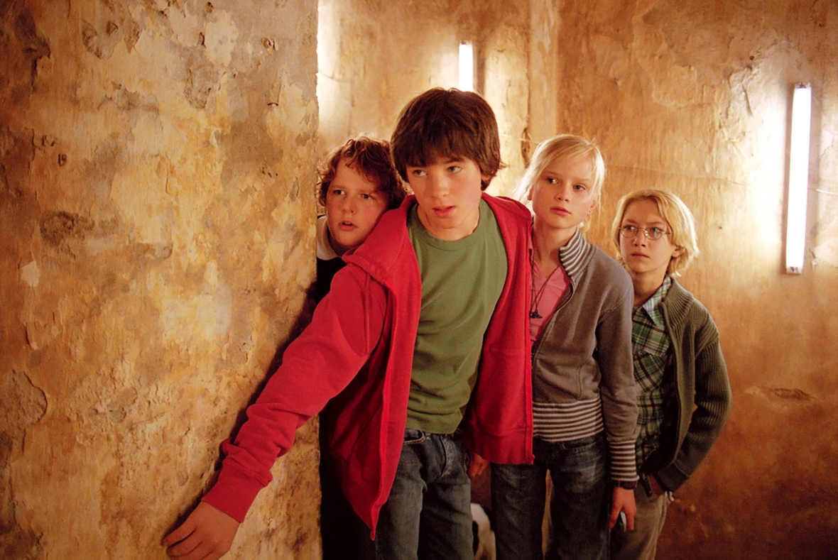 Szenenbild: Klößchen, Tim, Gaby und Karl, von links nach rechts im Bild, schleichen an der der Wand lang und verfolgen die Entführer