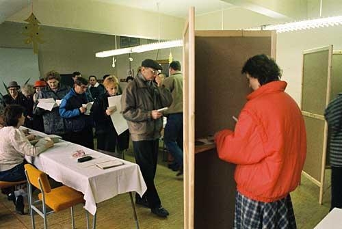 Die erste gesamtdeutsche Bundestagswahl fand am 2. Dezember 1990. Im Wahllokal befinden sich zahlreiche Menschen, eine Frau steht in einer Wahlkabine und wählt.