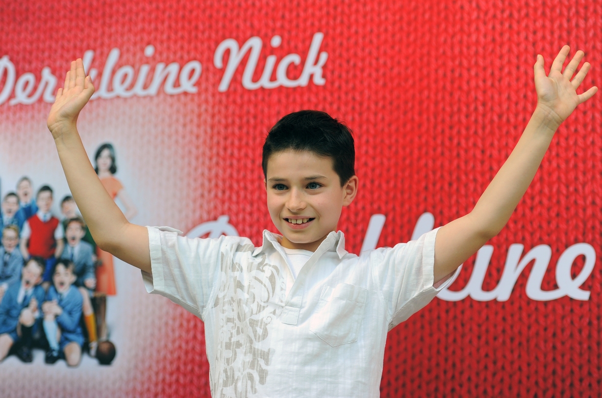 Maxime Godart, der den kleinen Nick spielt, strahlend und mit erhobenen Händen vor dem Filmplakat