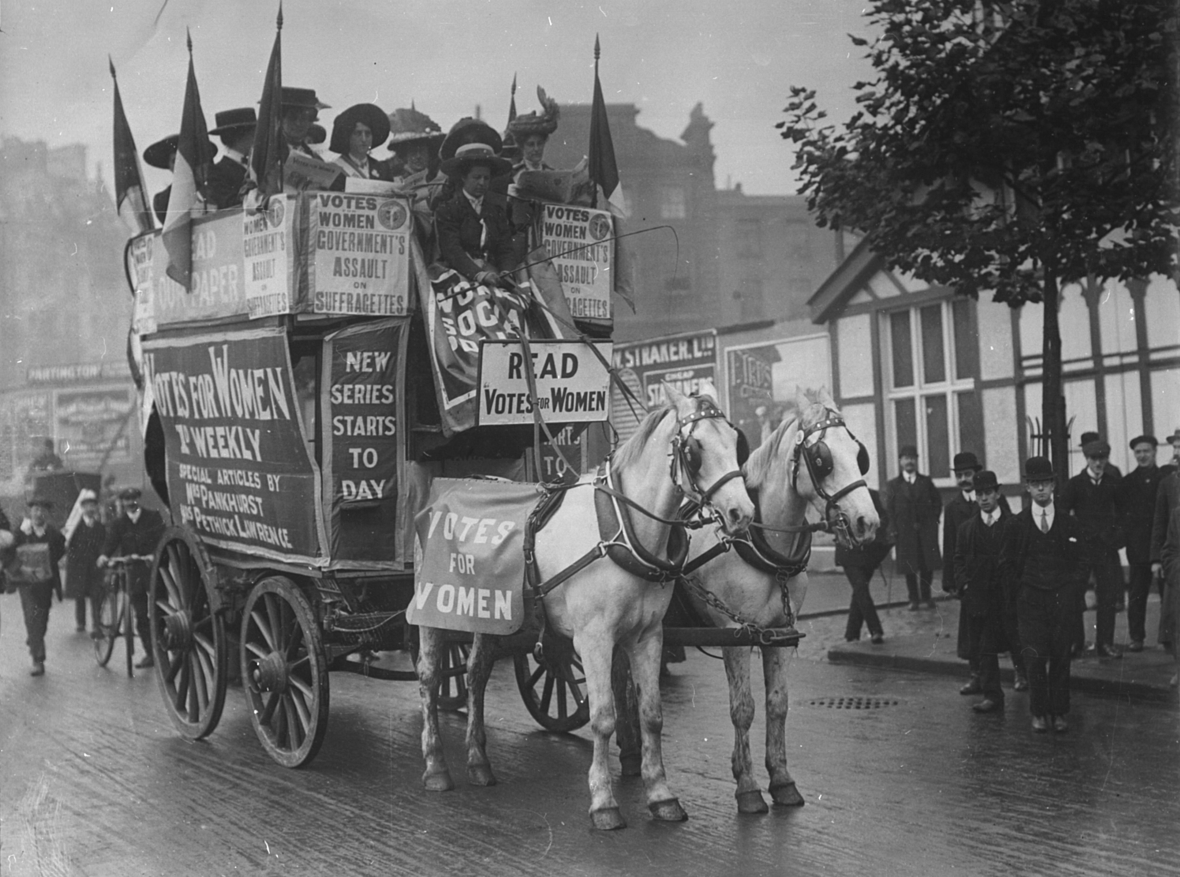 Demonstration von Suffragetten um 1905. Man sieht eine Kutsche mit Aktivistinnen und vielen Plakaten, auf denen das Wahlrecht für Frauen gefordert wird.