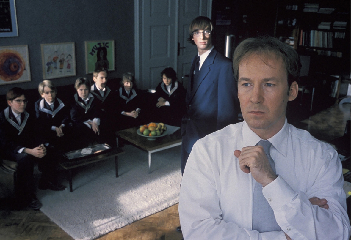 Szenenbild: Der Lehrer Justus Bökh, rechts im Bild, schaut nachdenklich und Schüler im Hintergrund blicken erwartungsvoll auf ihn 