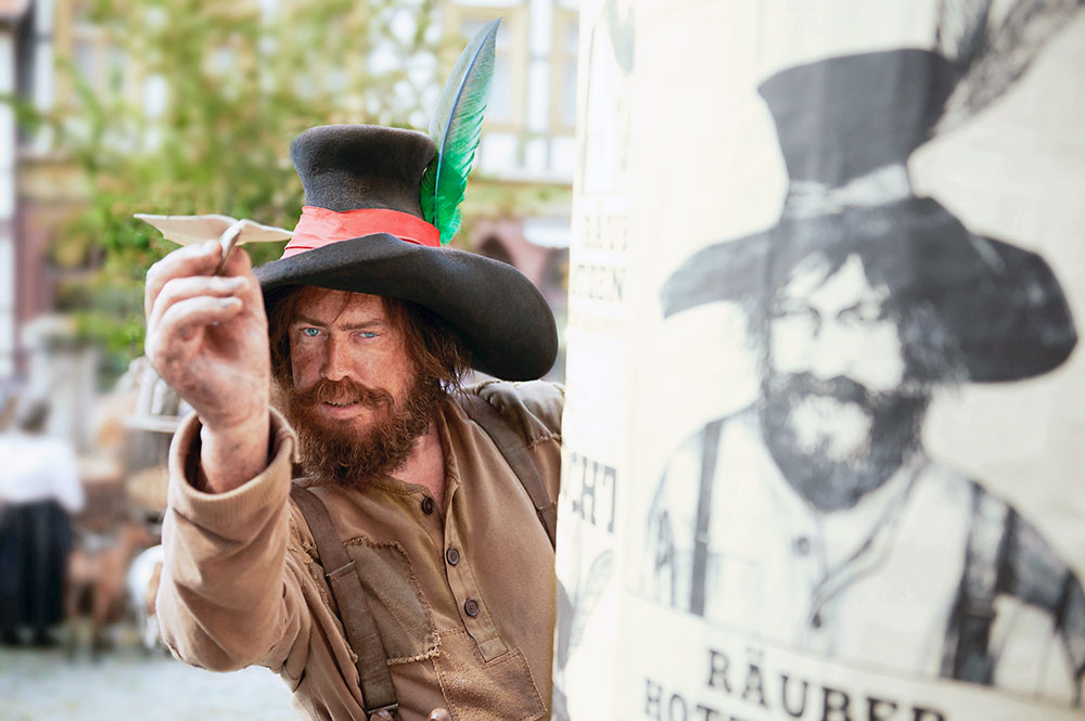 Szenenbild: Der Räuber Hotzenplotz mit Bart und einem großen Hut hat in der rechten Hand einen Papiervogel. Er hat darauf eine Nachricht geschrieben, die er versendet.