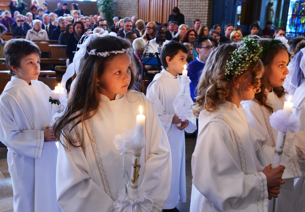 Katholische Mädchen und Jungen feiern das Fest ihrer Ersten Heiligen Kommunion. Die Kinder sind weiß gekleidet, tragen Kerzen und ziehen in die Kirche ein.