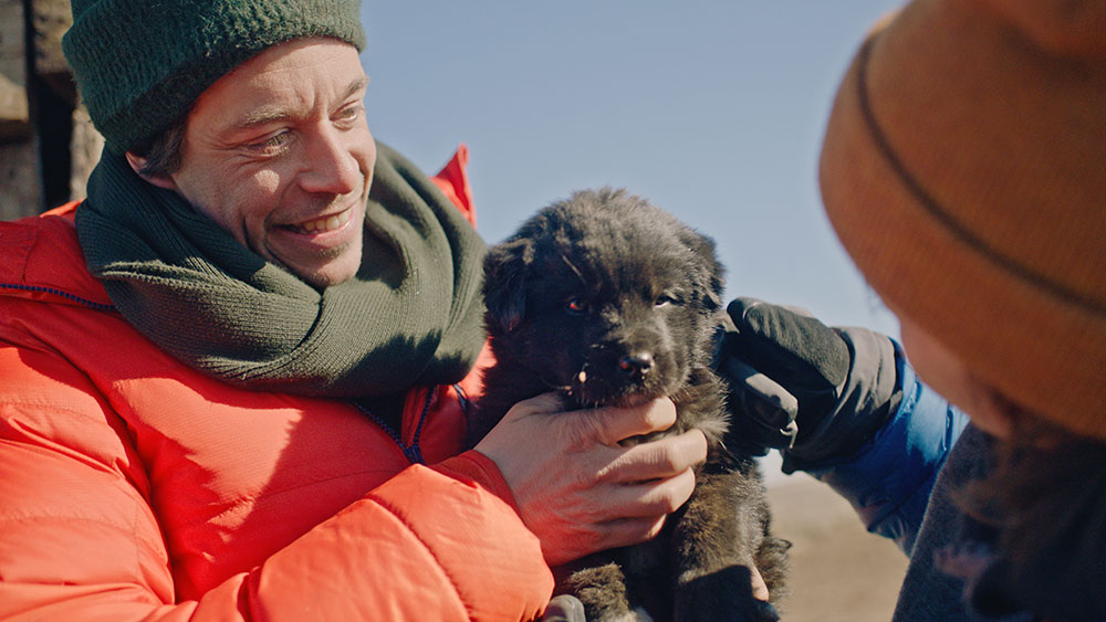 Szenenbild: Checker Tobi (links im Bild) mit Hundebaby in der Wüste Gobi. Er trägt eine rote Jacke und hat eine Wollmütze um.