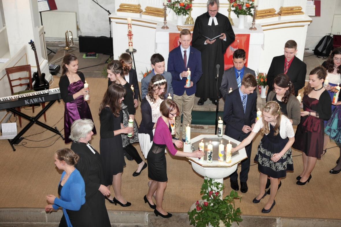 Junge evangelische Christen feiern ihre Konfirmation. Sie sind um ein Taufbecken in der Kirche gruppiert und haben zum Teil Kerzen in der Hand. 