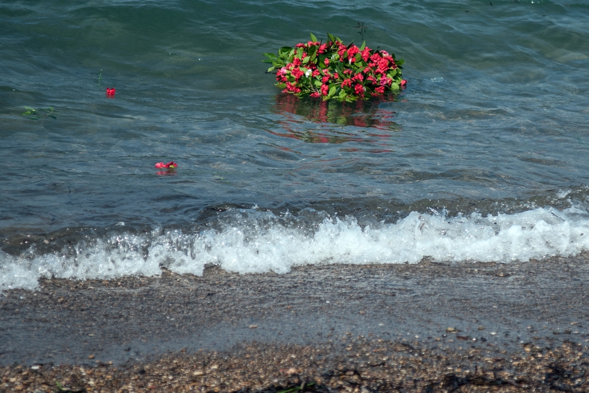 Ein Kranz mit Blumen schwimmt auf Meereswellen. Der Kranz erinnert an die vielen Menschen, die während ihrer Flucht über das Mittelmeer ertrunken sind.