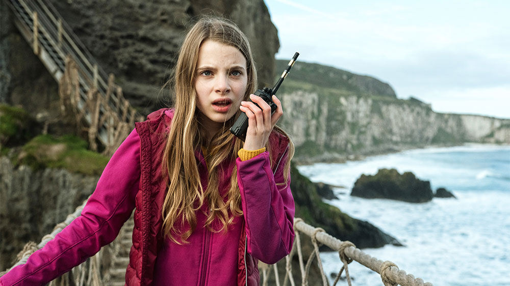 Szenenbild: Alice spricht in ein Walkie Talkie, das sie in der linken Hand hält. Im Hintergrund rechts sind die nordirischen Steilküste und das Meer zu sehen. Sie verfolgt den Dieb.