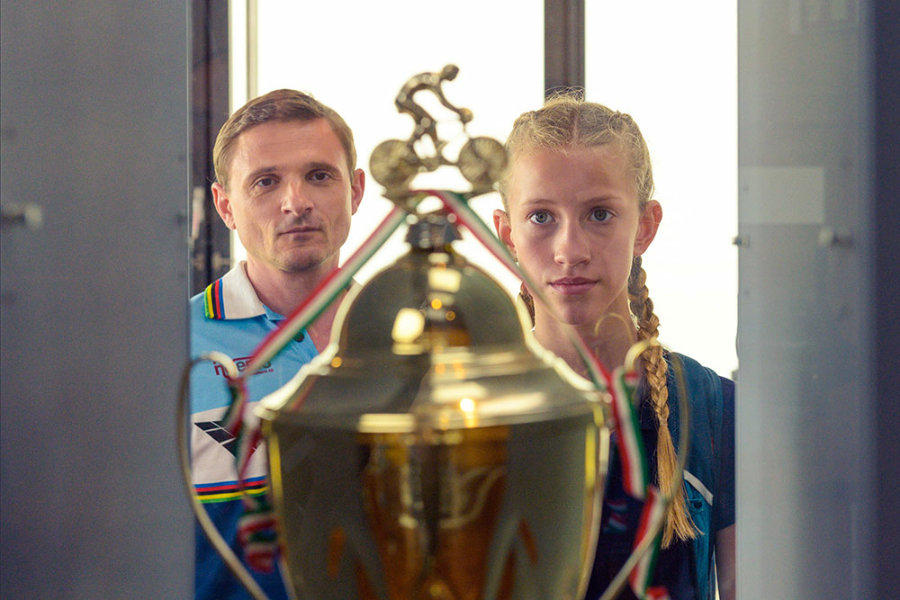 Szenenbild: Im Vordergrund des Bildes ist ein goldfarbener Siegerpokal. Dahinter stehen Madison (rechts im Bild) und ihr Vater (links im Bild) und bestaunen den Pokal.
