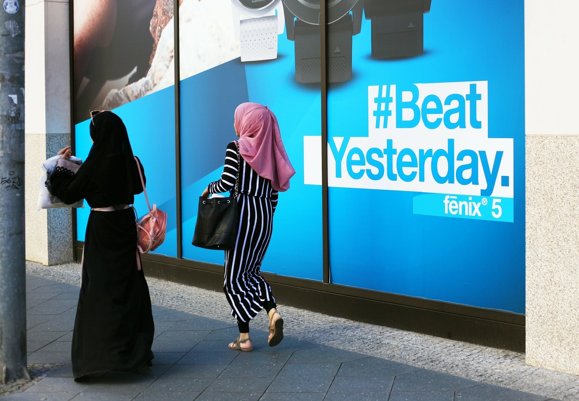 Zwei muslimische Frauen von hinten beim Passieren eines Plakats. Eine Frau trägt ein langes schwarzes Gewand, die andere ist modern gekleidet.