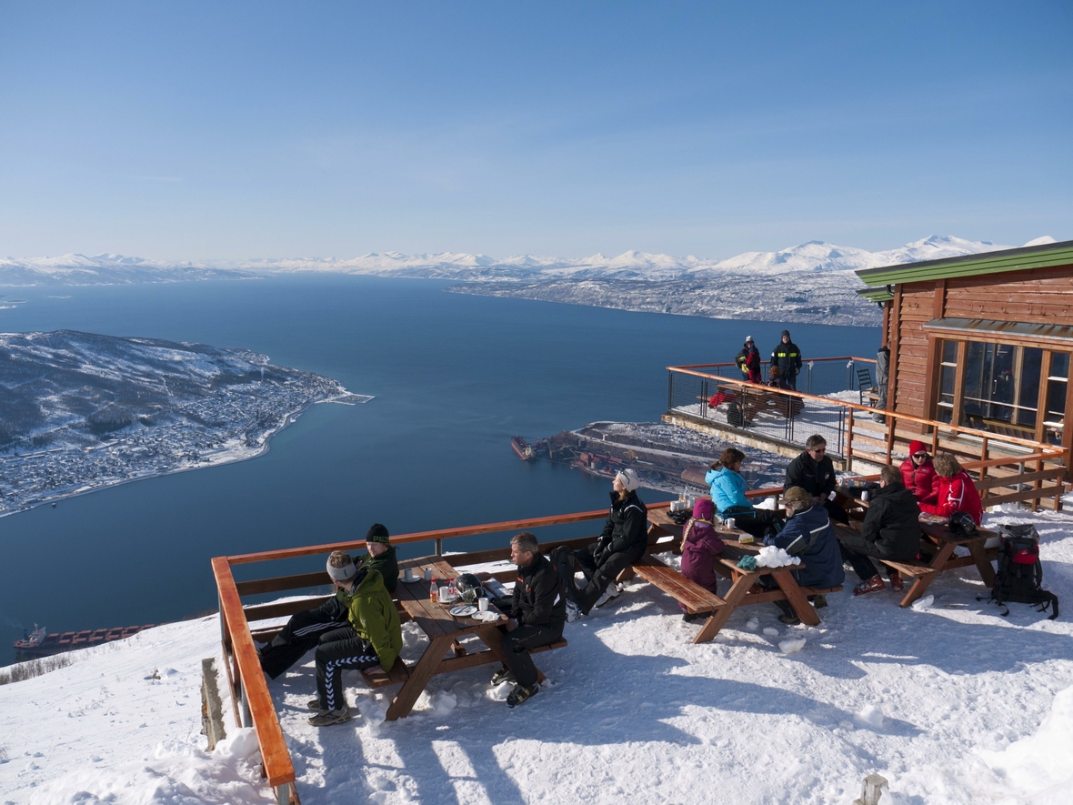 Wintersportler sitzen hoch über dem Ofotfjord am Berg Fagernes-Fjellet im Schnee und genießen das sonnige Wetter im norwegischen Narvik.
