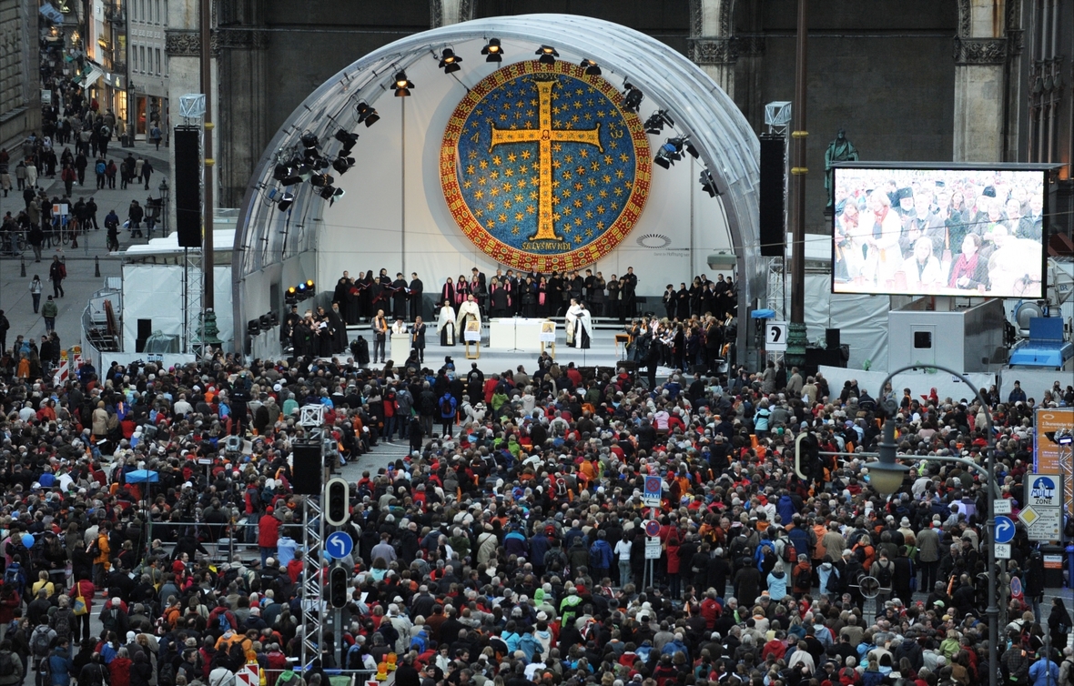 Christen und Christinnen treffen sich beim gemeinsamen Kirchentag in München 2010. Viele Menschen versammeln sich vor einem großen Zelt, in dem ein großes Kreuz den sichtbaren Hintergrund bildet. 