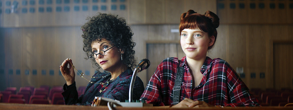 Szenenbild:  Idas Lehrerin Miss Cornfield mit Brille und lockigen schwarzen Haaren (links im Bild) sitzt neben Ida (rechts im Bild) mit roten hochgesteckten Haaren. Sie unterstützt Ida bei ihrer Regiearbeit.