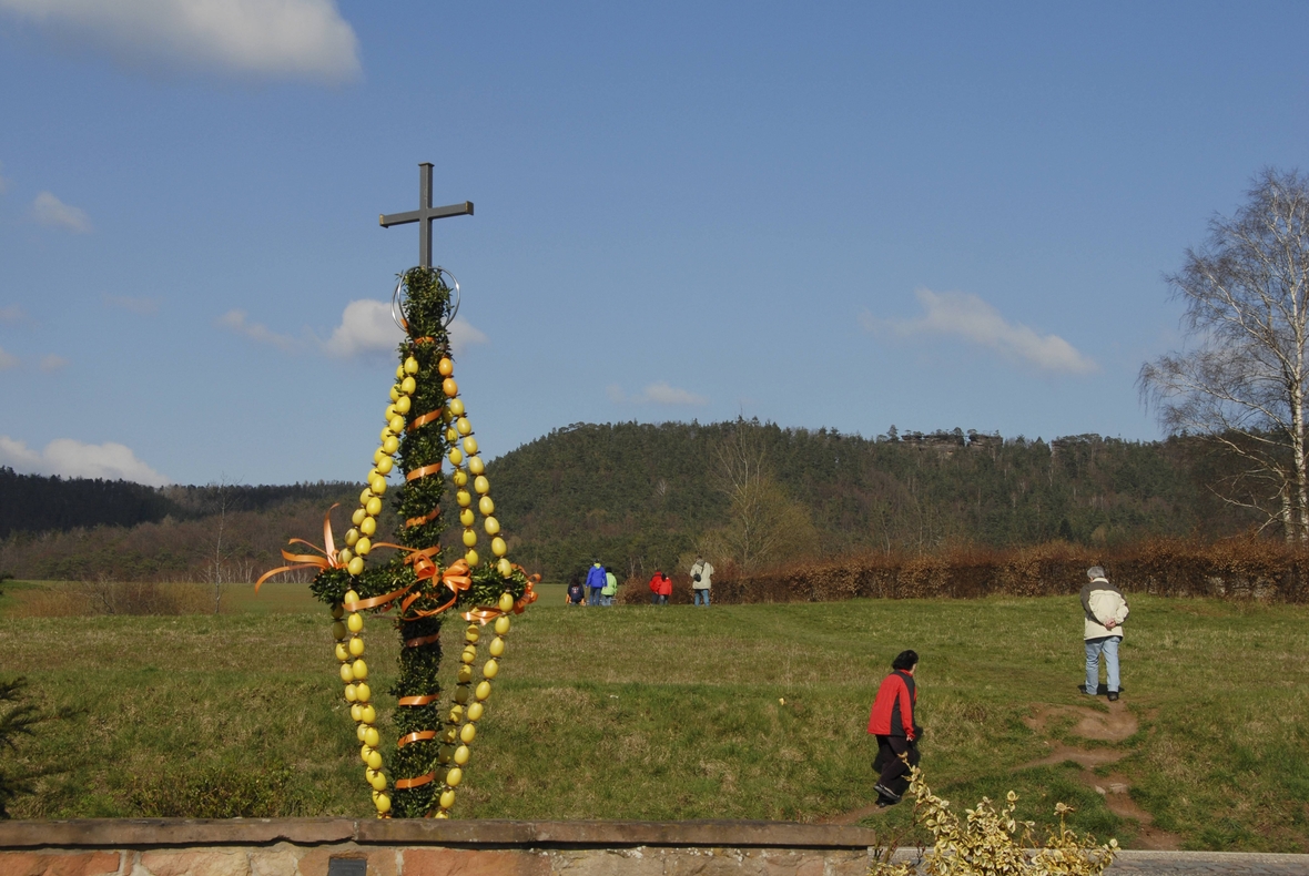 Ein geschmücktes Osterkreuz. Wanderer sieht man von hinten, die sich auf einer großen Wiese auf das Osterkreuz zubewegen.