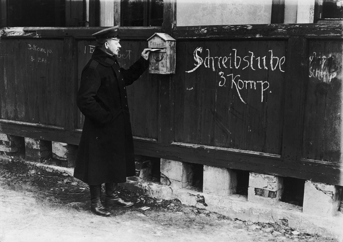 Ein Briefkasten ist an der Schreibstube 3. Kompagnie im 1. Weltkrieg angebracht. Ein Mensch mit Uniform wirft einen Brief in den Kasten.