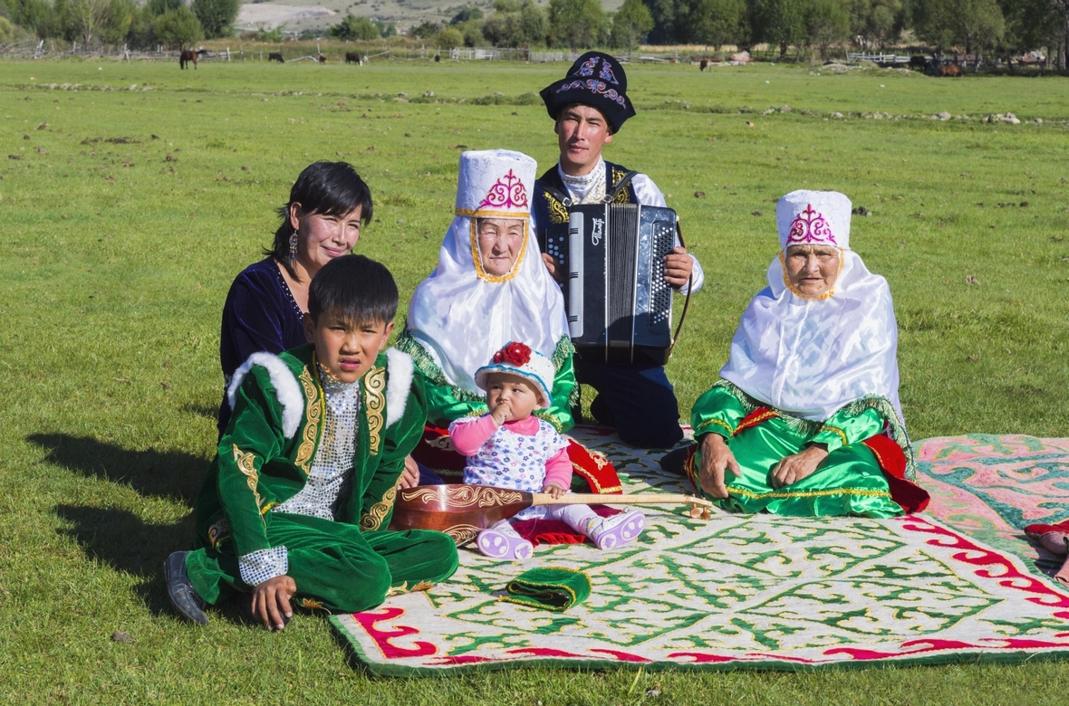 Kasachische Familie in traditioneller Kleidung hört der Musik eines Akkordeonspielers im Tian Shan Hochgebirge zu.

