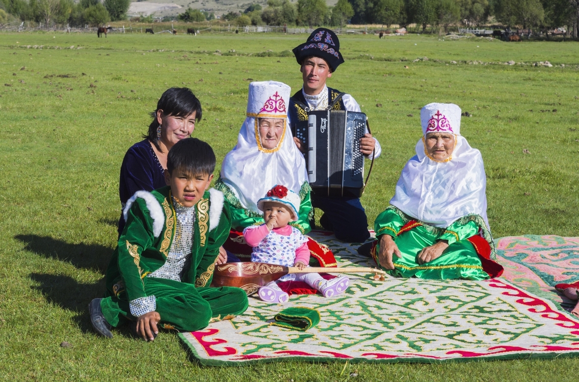 Kasachische Familie in traditioneller Kleidung hört der Musik eines Akkordeonspielers im Tian Shan Hochgebirge zu.

