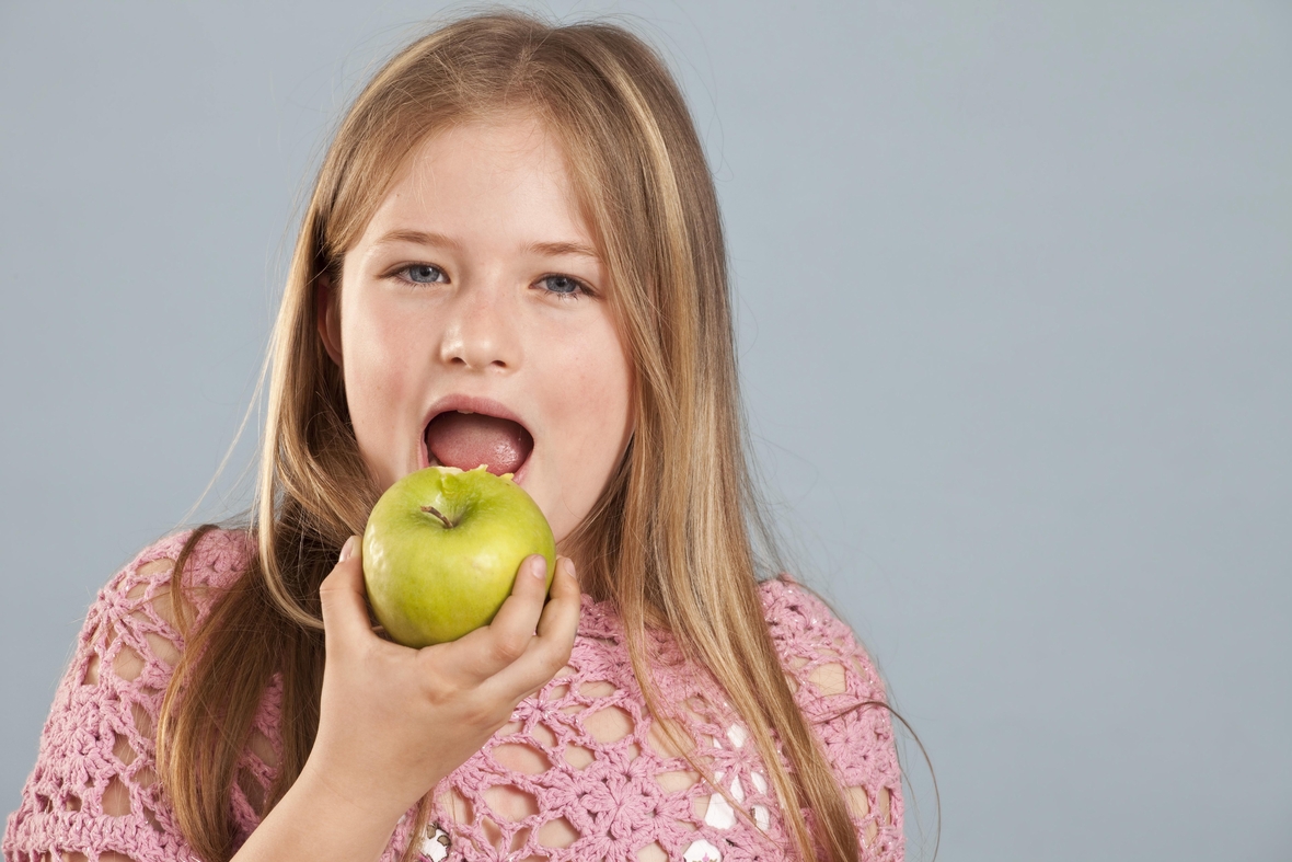 Ein Mädchen isst einen Apfel. Sie hält den Apfel in der Hand und öffnet zum Abbeißen ihren Mund.