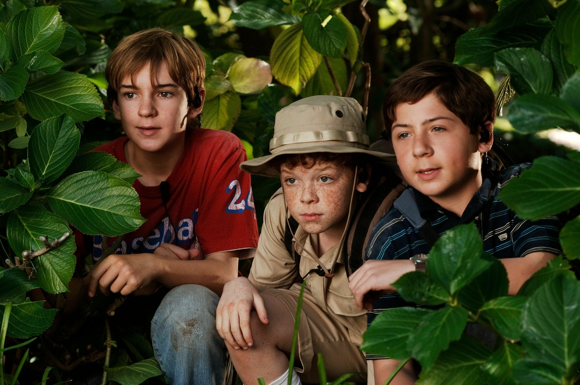 Szenenbild: Die drei jungen Detektive hocken im Gebüsch und beobachten heimlich etwas.