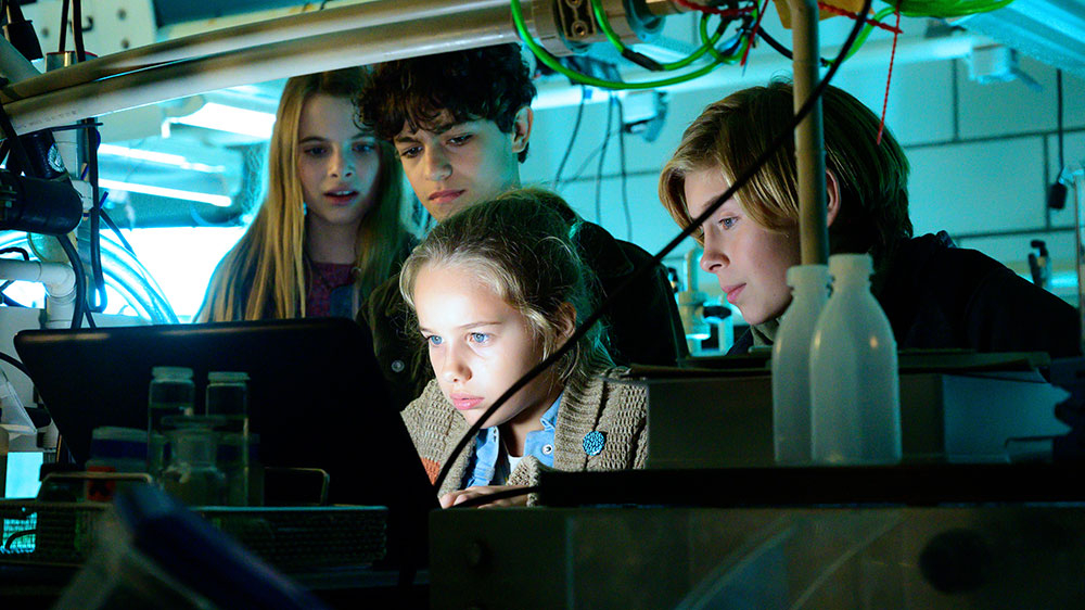 Szenenbild: Clarissa sitzt am Computer im Forschungslabor und hat etwas entdeckt. Hinter ihr stehen Alice, Tarun und Johnny (von links nach rechts.