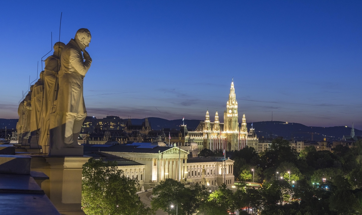 Das Foto zeigt einen Ausblick bei Nacht auf das erleuchtete Wien. Zu sehen sind das Parlament und Rathaus und links im Vordergrund die Statuen berühmter Wissenschaftler auf dem Dach des Naturhistorischen Museums.