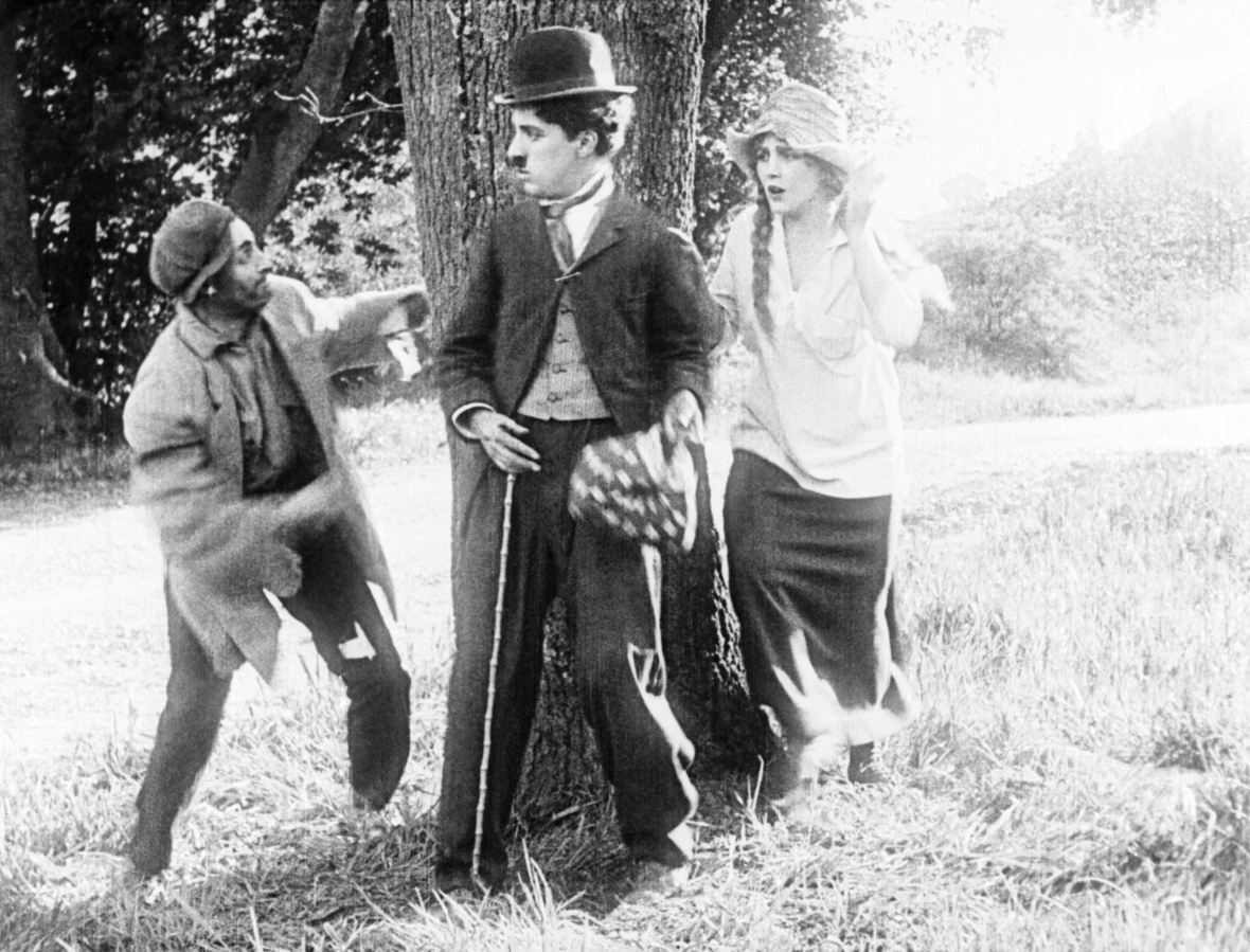 Das schwarz-weiß Bild zeigt Chaplin in einer seiner berühmtesten Rolle als der "Tramp". 1915. Neben ihm sind die Schauspieler Leo White und Edna Purviance in ihren Filmrollen zu sehen.