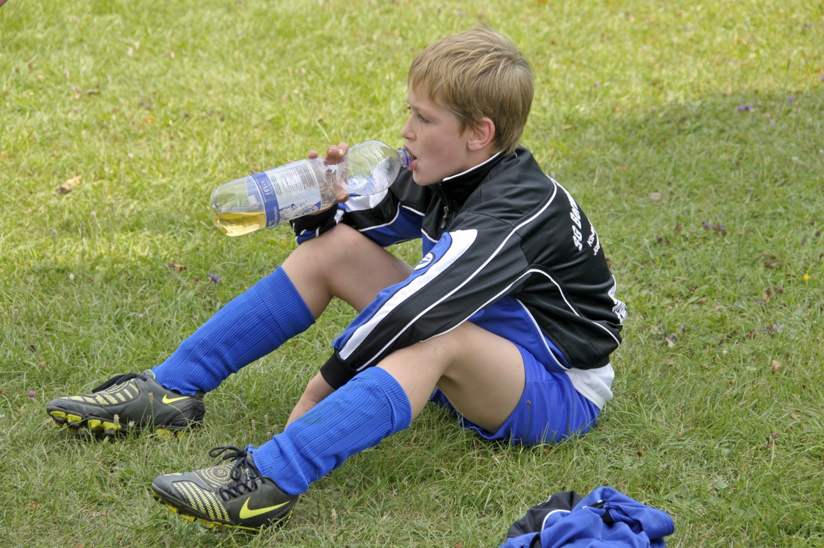 Ein  Junge sitzt auf dem Rasen und trinkt nach dem Fußball-Spiel Wasser aus einer Flasche, denn beim Sport verbraucht man viel Energie und Wasser. 