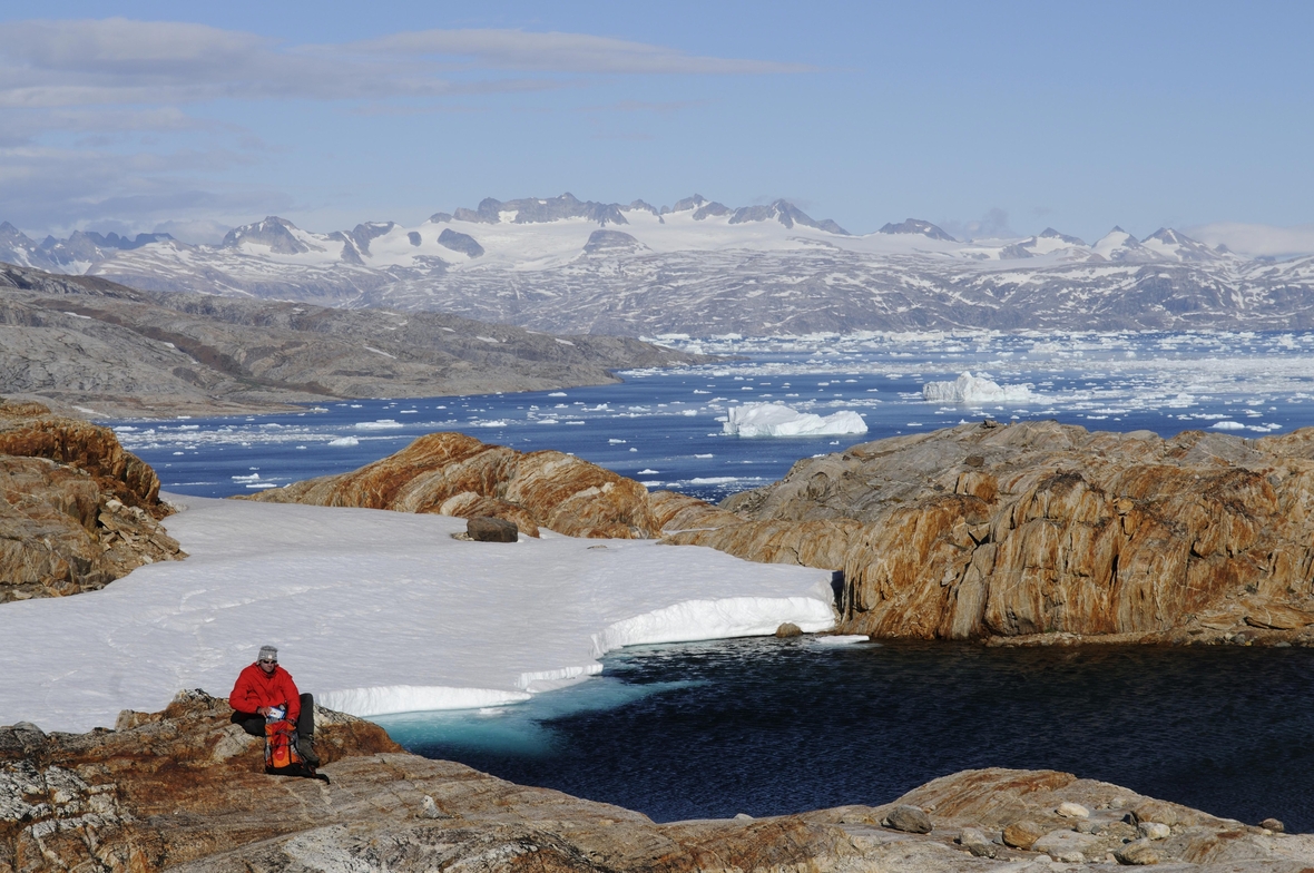Das Bild zeigt eisbedeckte Berge am Johan-Petersen-Fjord in Ostgrönland. Ein Wanderer macht gerade eine Pause beim Trekking durch die Eislandschaft. 