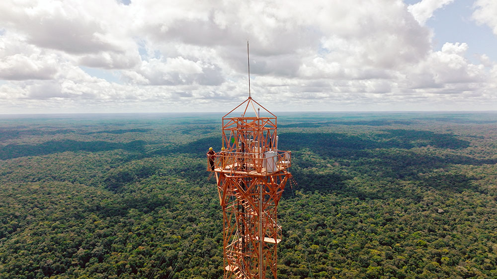 Szenenbild: Checker Tobi mit Marina auf dem Atto-Turm im brasilianischen Urwald. Die beiden sind kaum zu erkennen. Man sieht den riesigen Urwald und den roten Stahlturm.
