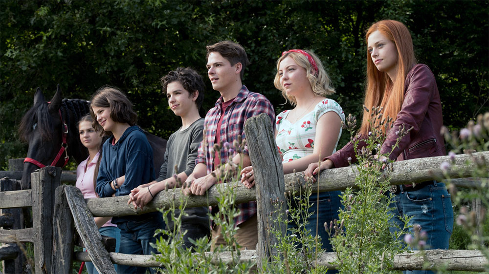 Szenenbild: Ari (zweite von links im Bild) und Mika (rechts außen im Bild) stehen mit ihren Freunden an der Pferdekoppel. Sie stützen sich auf dem Geländer auf.dem Ge