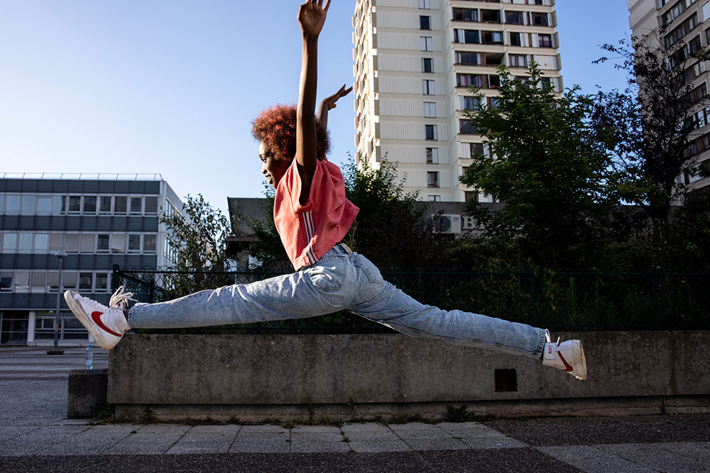 Szenenbild: Neneh in Jeans und T-Shirt macht vor Freude einen Spagat-Luftsprung. Im Hintergrund sind Häuser zu sehen.