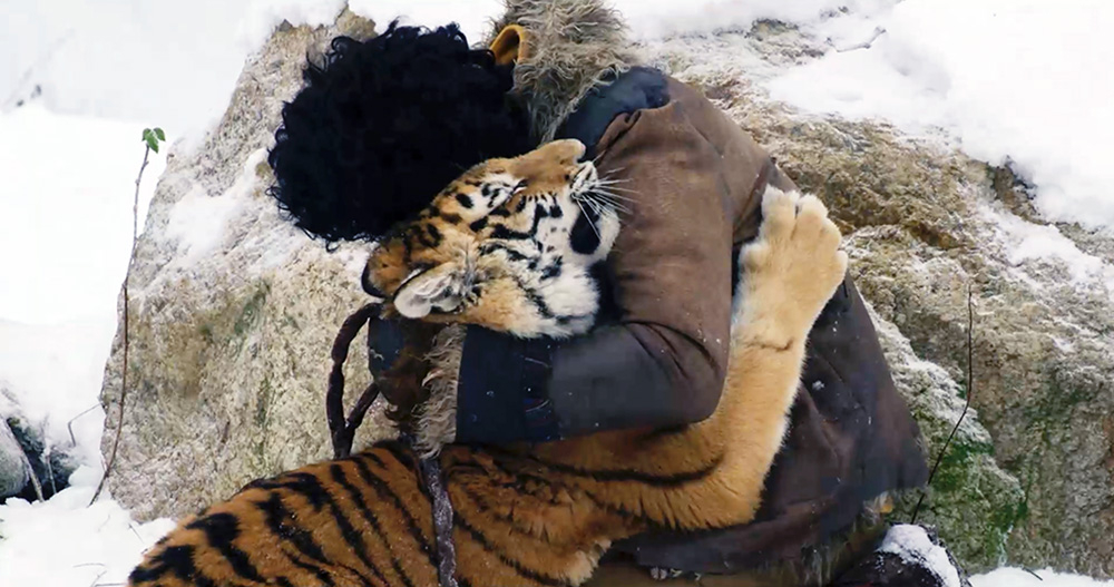 Szenenbild: Der Junge Balmani und das Tigerbaby Mukti umarmen sich. Sie sind in einer Schneelandschaft und hinter ihnen sind Berge zu sehen.