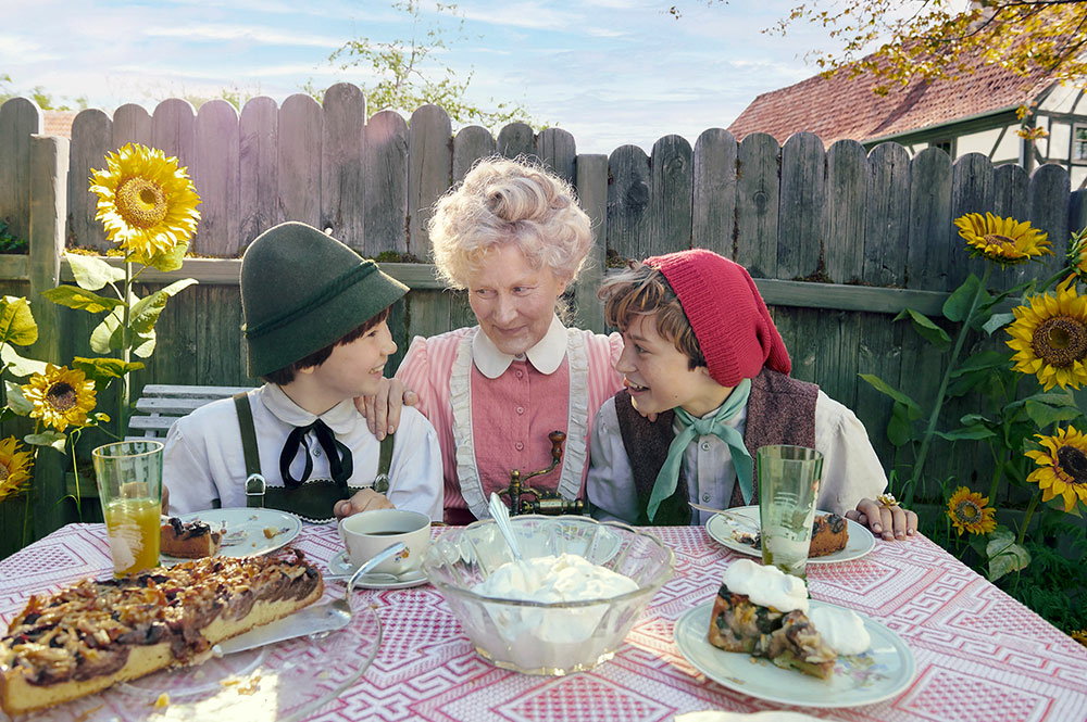 Szenenbild: Die Großmutter sitzt zwischen Seppel (links) und Kasperl (rechts) an einem Tisch im Garten.