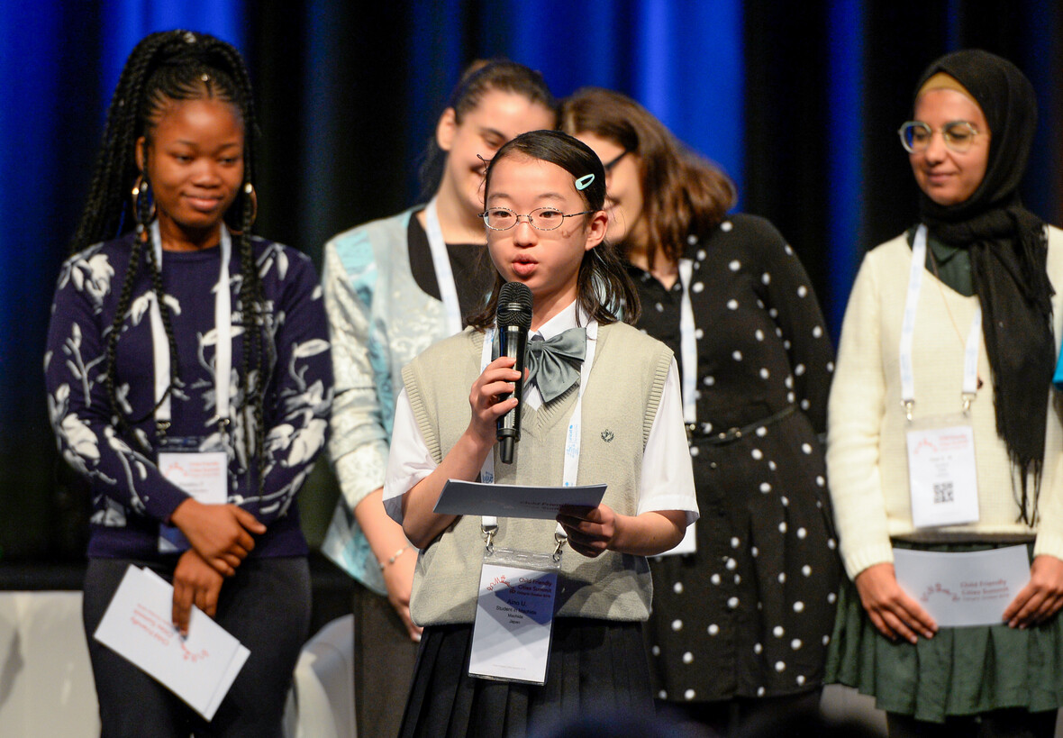 Ein Mädchen spricht auf einer Bühne ins Mikrofon, andere Kinder im Hintergrund.