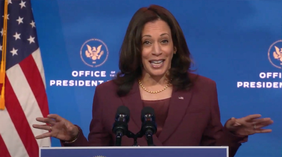 Kamala Harris, die erste weibliche Vizepräsidentin der USA. Sie spricht, neben der USA-Fahne stehend.