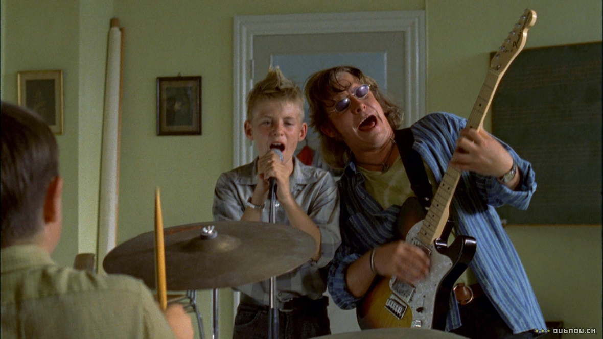 Szenenbild: Frits (links) mit einem Mikrofon in der Hand und sein neuer Lehrer Freddie (rechts) mit Gitarre spielen Rock’n’Roll. Beide singen dazu.