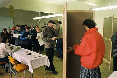 Die erste gesamtdeutsche Bundestagswahl fand am 2. Dezember 1990. Im Wahllokal befinden sich zahlreiche Menschen, eine Frau steht in einer Wahlkabine und wählt.