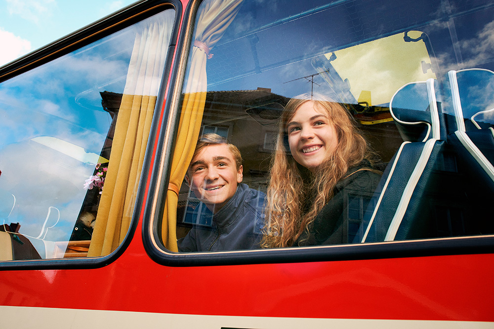 Szenenbild: Alfons (links) und Leonie (rechts) sitzen nebeneinander im roten Bus und schauen aus dem Fenster. Sie fahren auf Klassenfahrt.