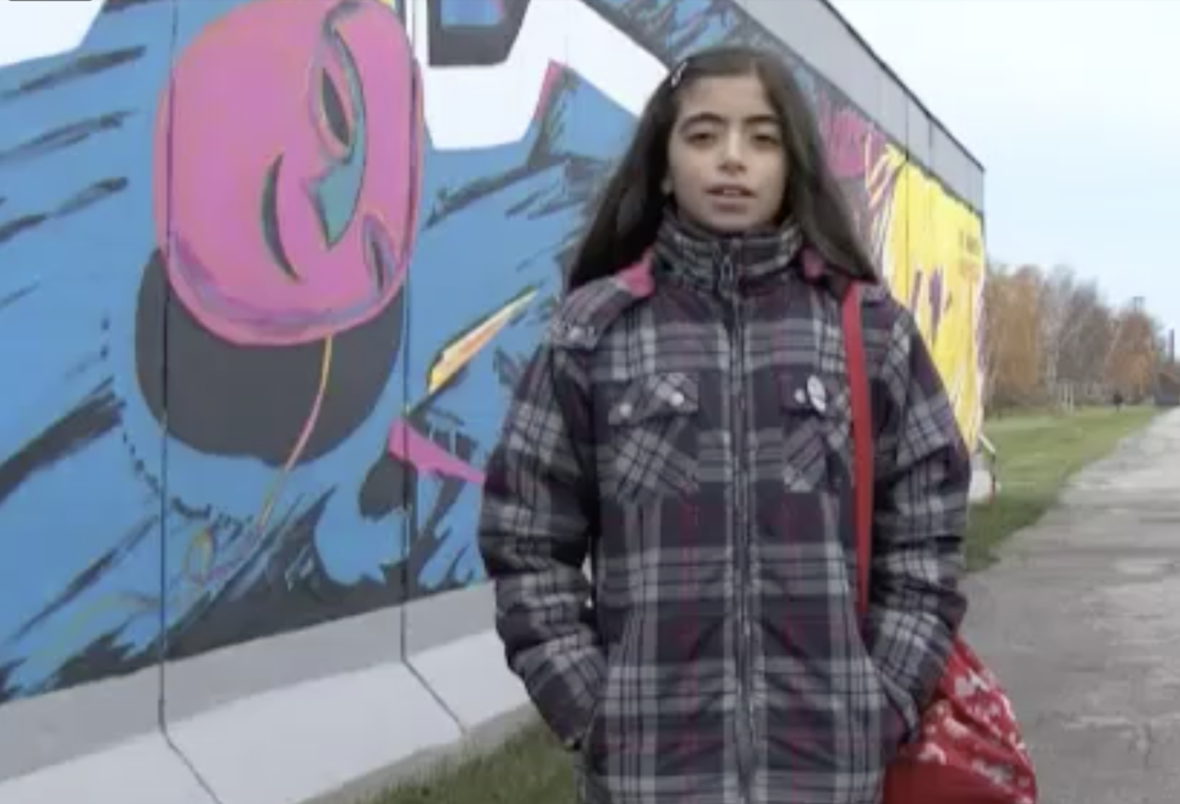 Ein Mädchen steht vor einem bemalten Teil der Berliner Mauer und beantwortet Fragen