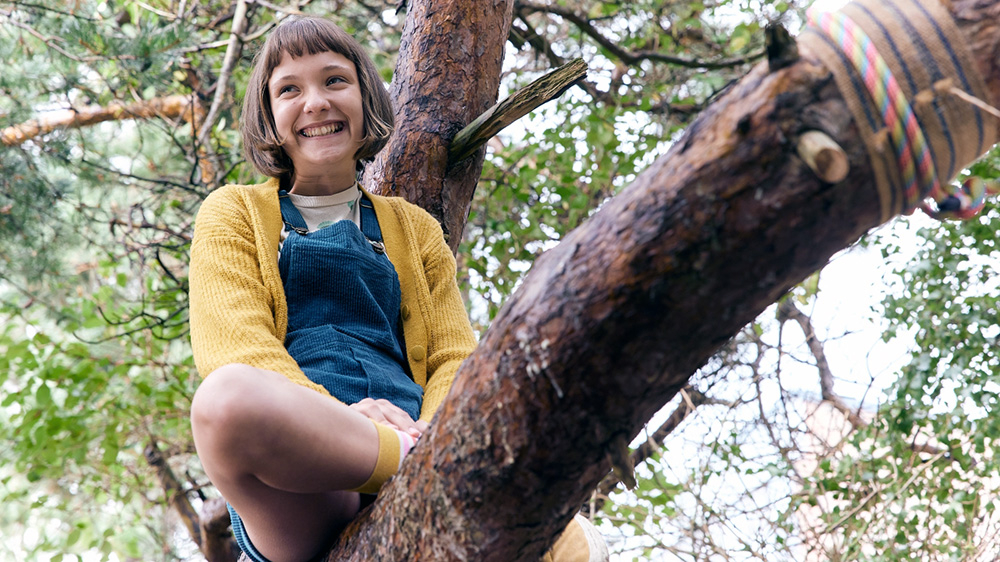 Szenenbild: Lotta sitzt lachend auf einem Baum. Sie hat eine "Prinz-Eisenherz"-Frisur und trägt eine gelbe Jacke.