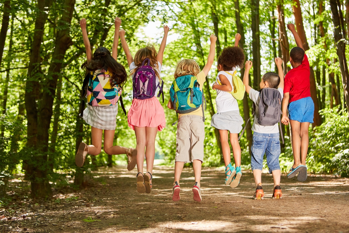 Kinder springen im Sommer im Wald in die Luft. Nicht nur am Weltspieltag macht das Spielen im Wald viel Spaß. 
