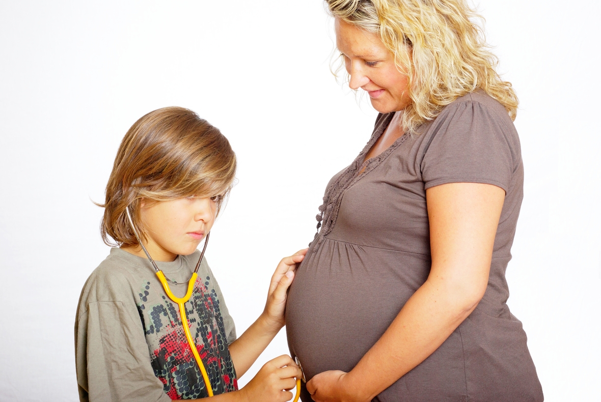 Eine schwangere Frau mit ihrem Sohn, der mit einem Stethoskop am Bauch seiner Mutter hört. 