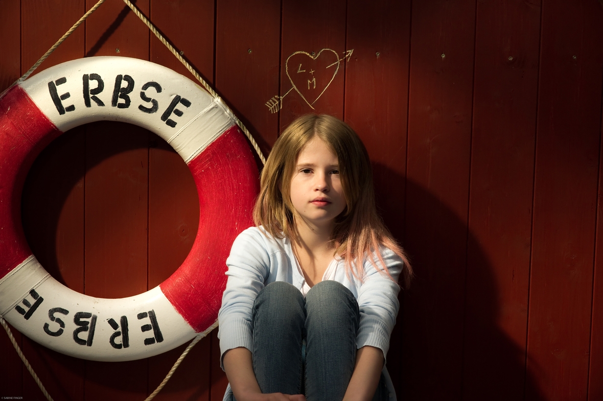 Szenenbild: Lola sitzt auf dem Hausboot Erbse, an der Wand hängt ein Rettungsring