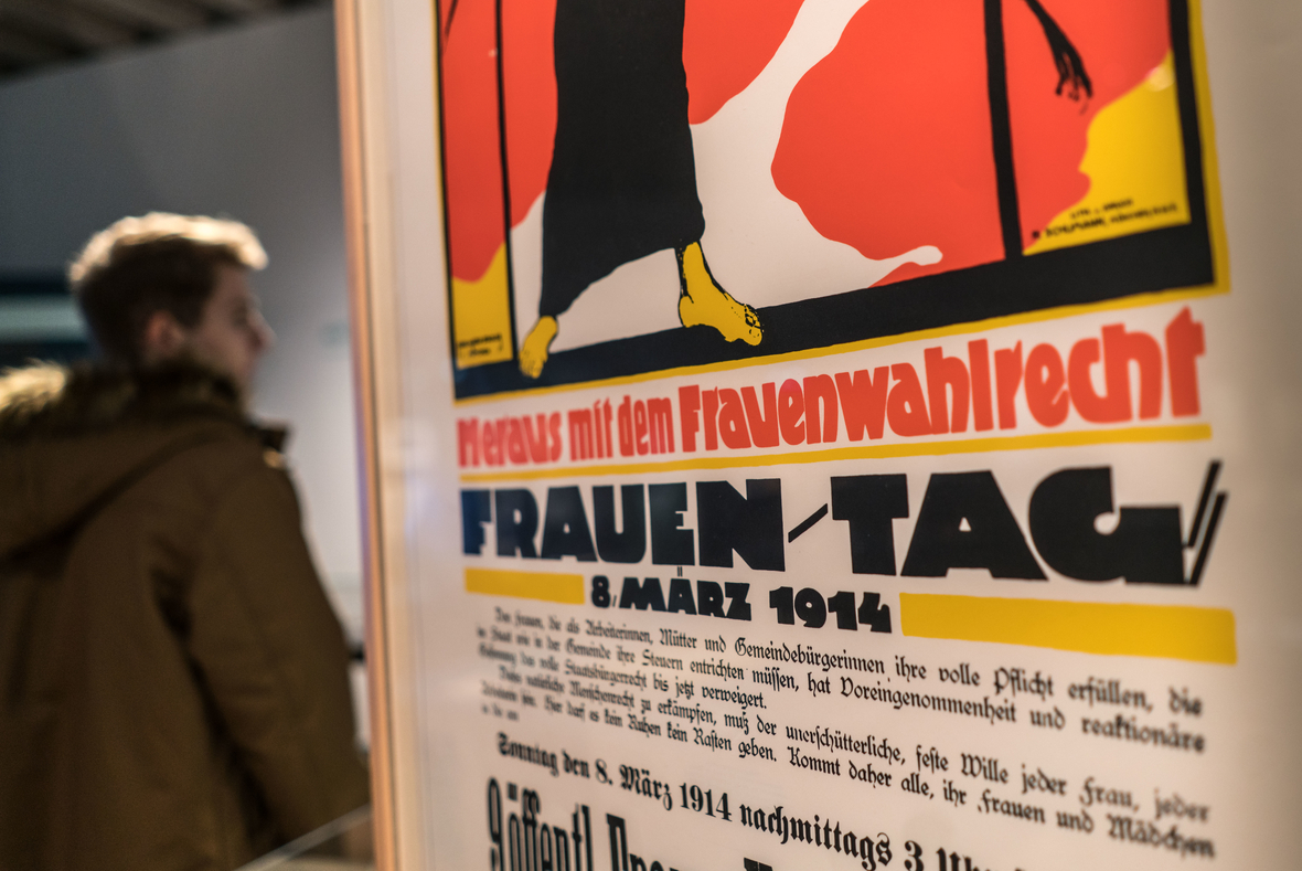 Ein Besucher der Ausstellung "Damenwahl" steht im Historischen Museum Frankfurt hinter einem Plakat zum Frauen-Tag 1914, auf dem "Heraus mit dem Frauenwahlrecht" gefordert wird.