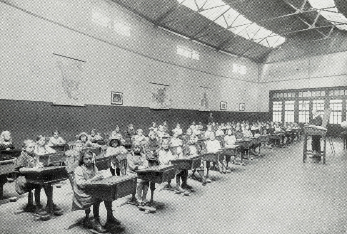1915: Englische Schulklasse während des Ersten Weltkriegs. Die Kinder sitzen an Einzelpulten. Vorne hält der Lehrer einen Vortrag.