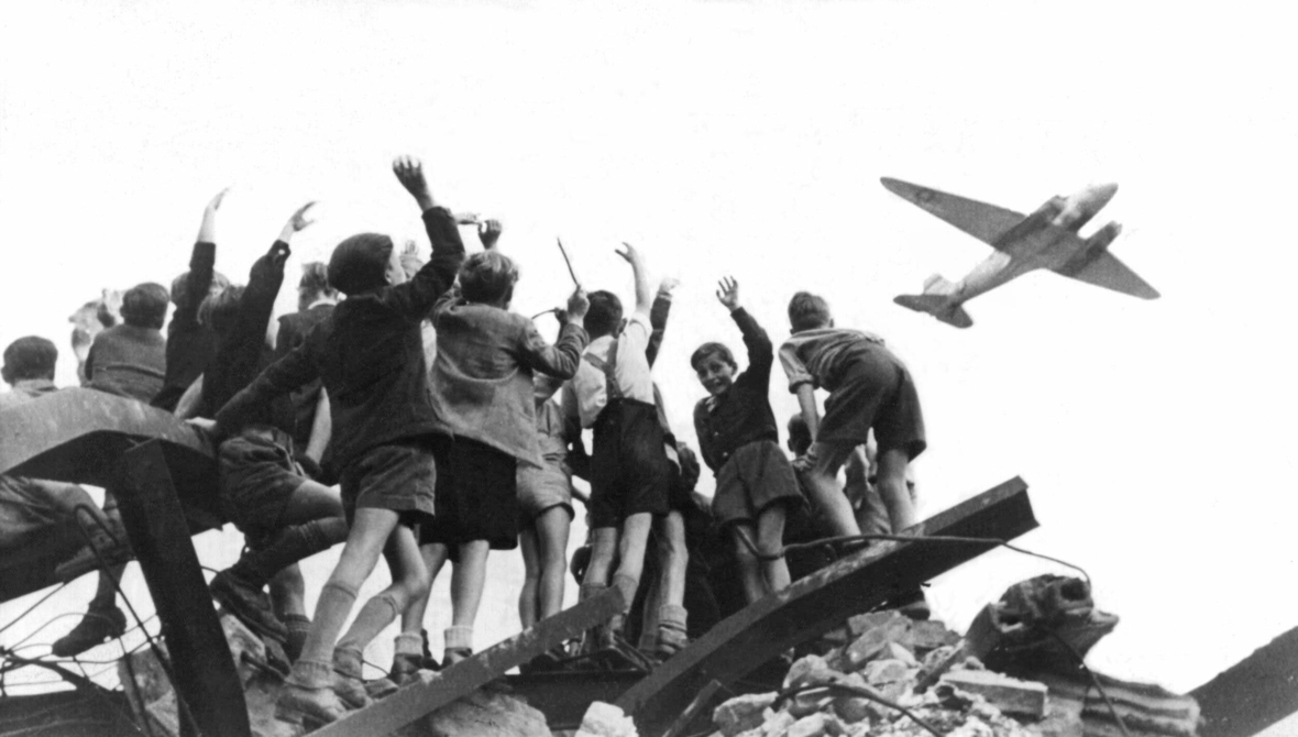 West-Berliner Kinder stehen auf einem Trümmerhaufen und begrüßen ein amerikanisches Transportflugzeug, das 1948 Versorgungsgüter nach West-Berlin bringt.