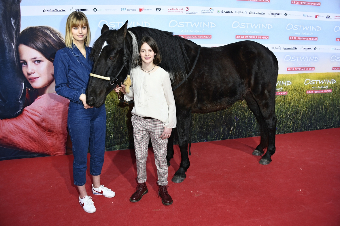 Links im Bild steht Hanna Binke, die MIka spielt, und rechts im Bild Luna Paiano, die Aris verkörpert. Sie halten das Pferd Ostwind in ihrer Mitte und stehen vor dem Filmplakat Ostwind - Aris Ankunft.