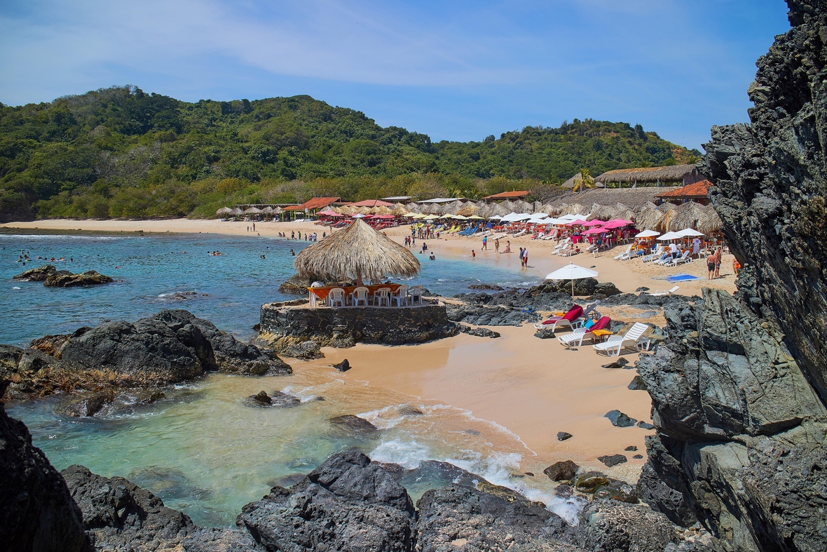 Ein Korallenstrand in Mexiko. Gezeigt wird eine Bucht am Meer mit Strand, Menschen und Hügeln im Hintergrund