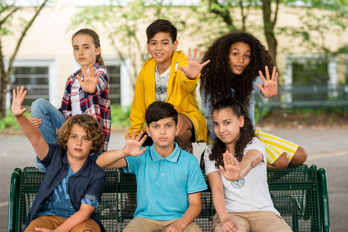Sechs Kinder posieren auf einer Bank für ein Gruppenbild und strecken eine Hand nach vorne aus.