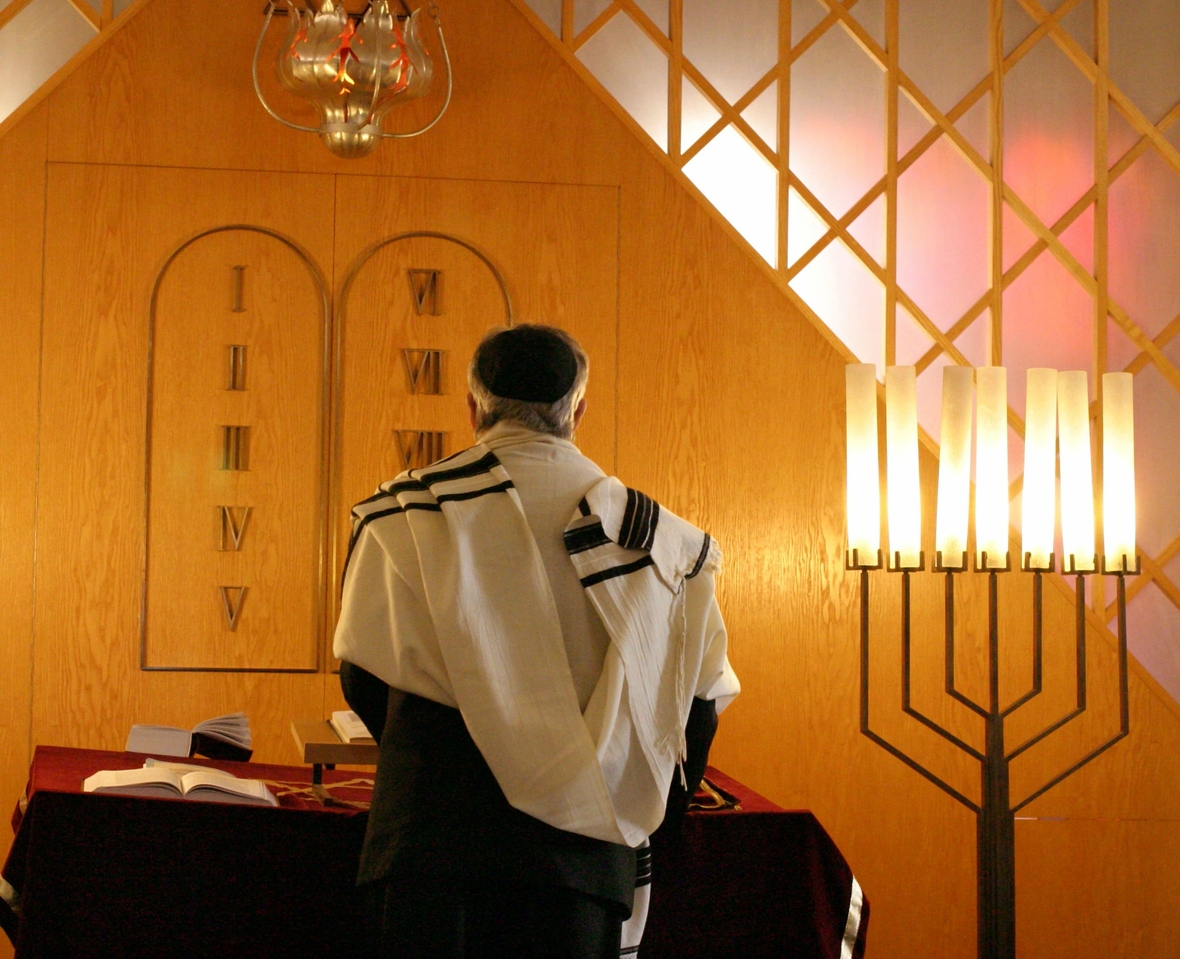 Ein Jude mit dem umgelegten weißen Tallit, dem Gebetstuch, beim Vorlesen während des Gottesdienstes. Daneben steht die Menora, der siebenarmige Leuchter. Sie ist für die Juden das Symbol für den Tempel, Gottes Gegenwart und das ewige Leben.