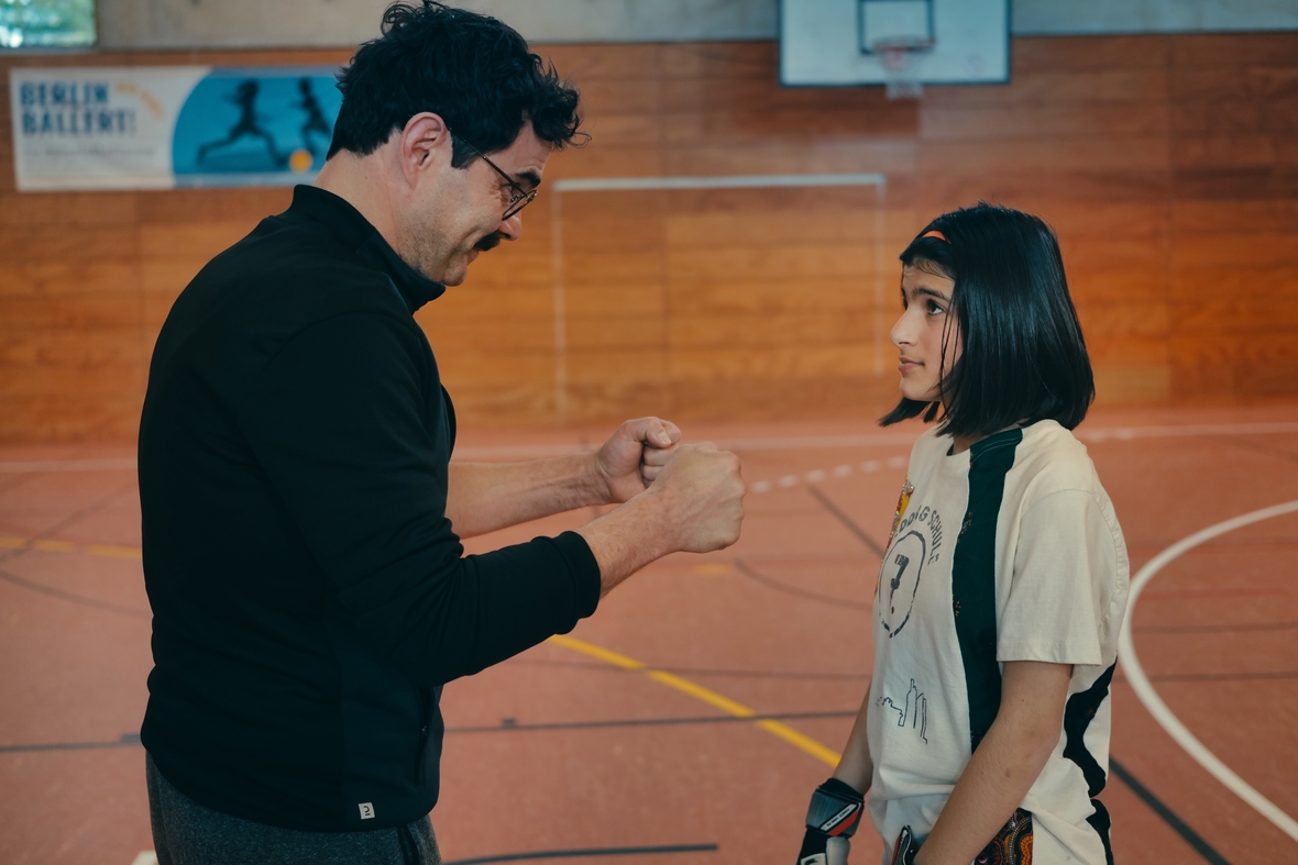 "Sieger Sein": Man sieht die Hauptfigur Mona und ihren Lehrer, Herr Che, in der Sporthalle, wie er Mona etwas erklärt.